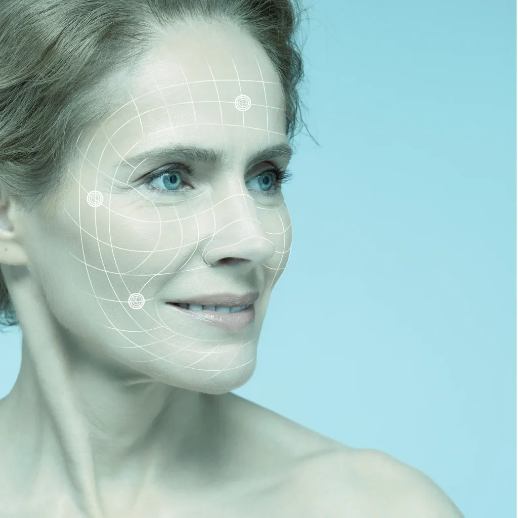A woman with fair skin and blue eyes looks to the side, smiling slightly. A digital grid overlay is mapped onto her face, suggesting a facial analysis development platform against a cool blue background.